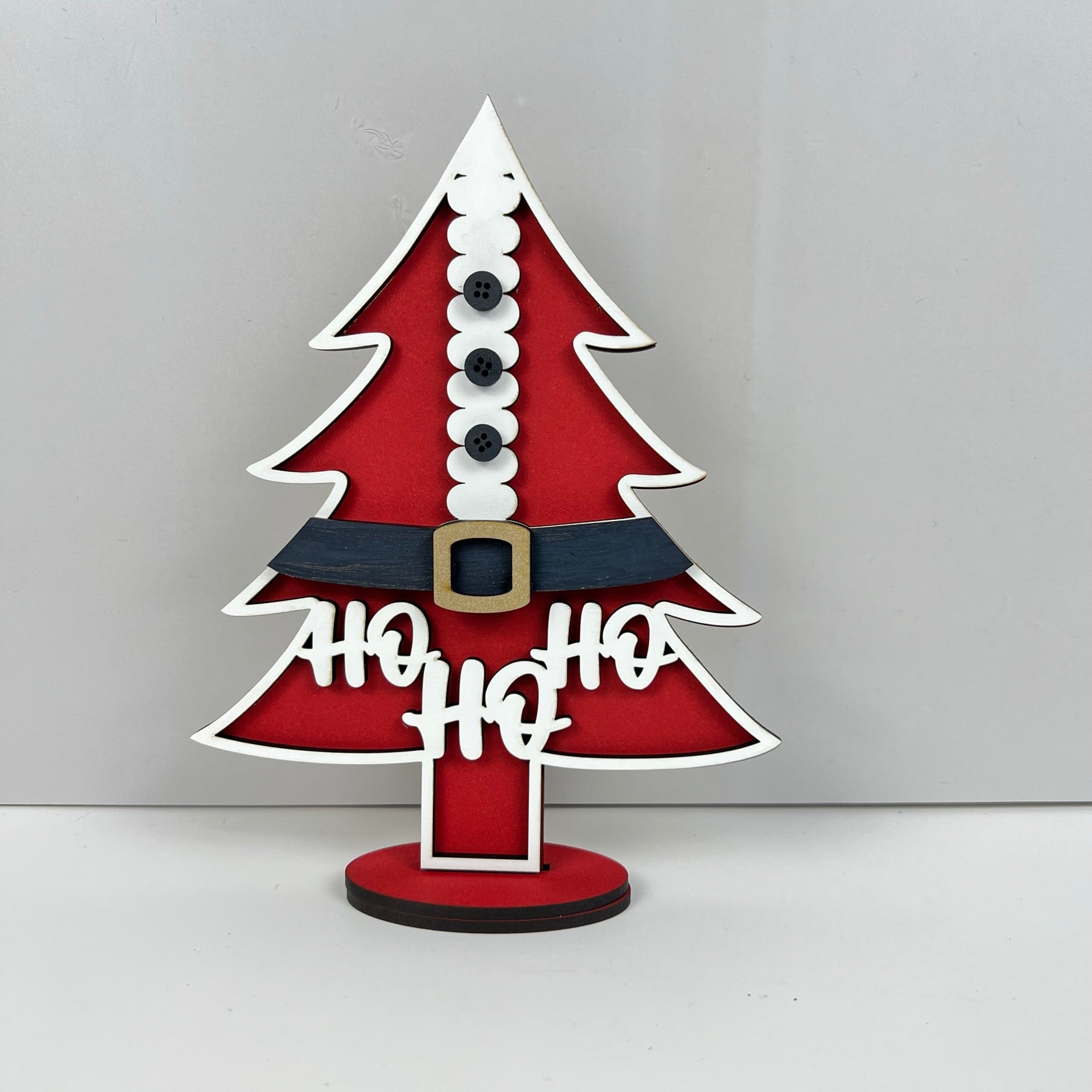 The Standing Christmas Tree Shelf Sitters from Janet's Craft Corner add festive charm to your holiday decor. Inspired by Santa's suit, they feature a red design with white trimming, black buttons, a black belt with a yellow buckle, and the words "Ho Ho Ho." Perfect for enhancing your shelf display this season.