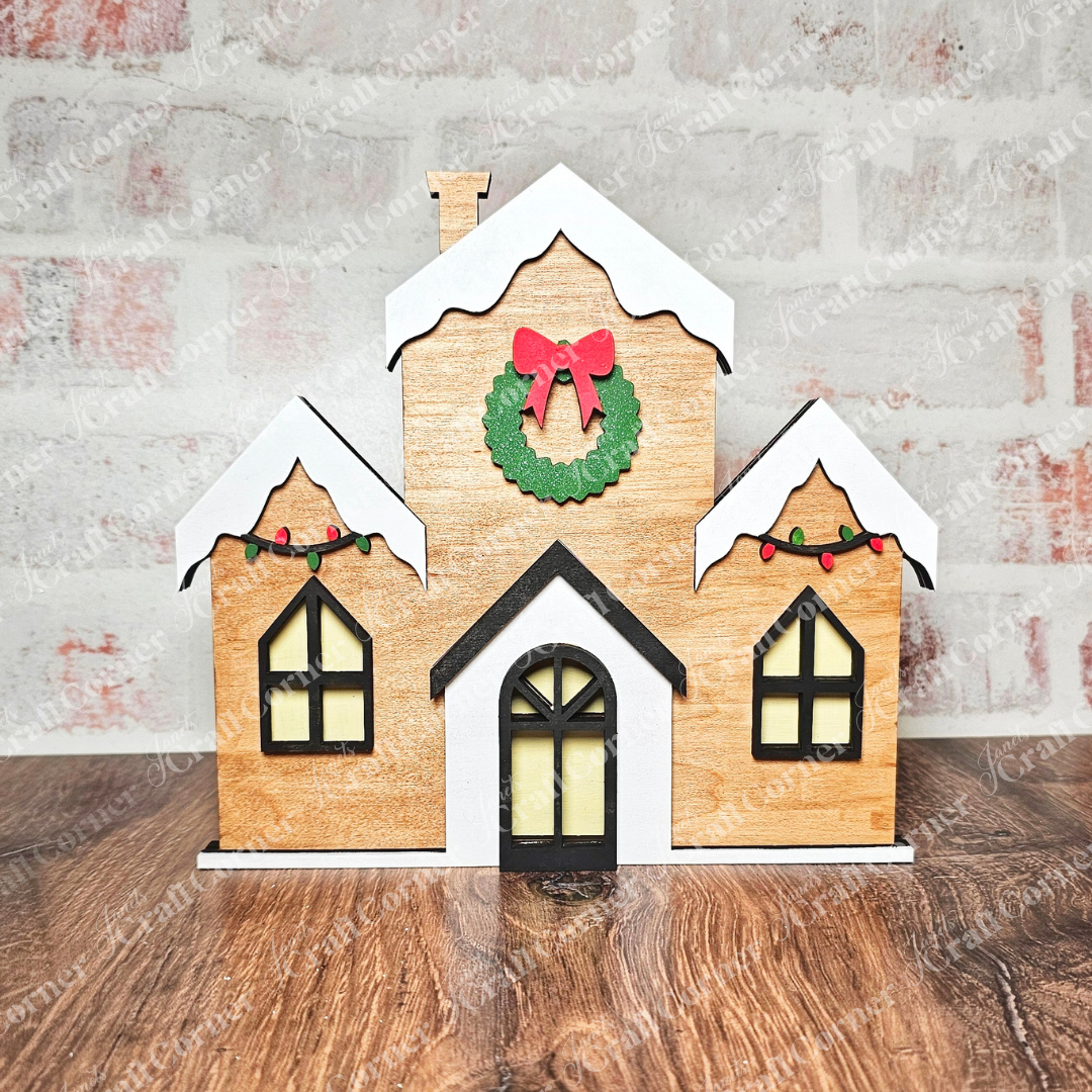 The Christmas Village Shelf Sitters - Set of 4 by Janet's Craft Corner encapsulates the holiday charm with a decorative wooden house adorned with a festive wreath and bow, flanked by two smaller sections featuring snow-like roofs and string lights. The ensemble is beautifully set against a textured brick wall backdrop.