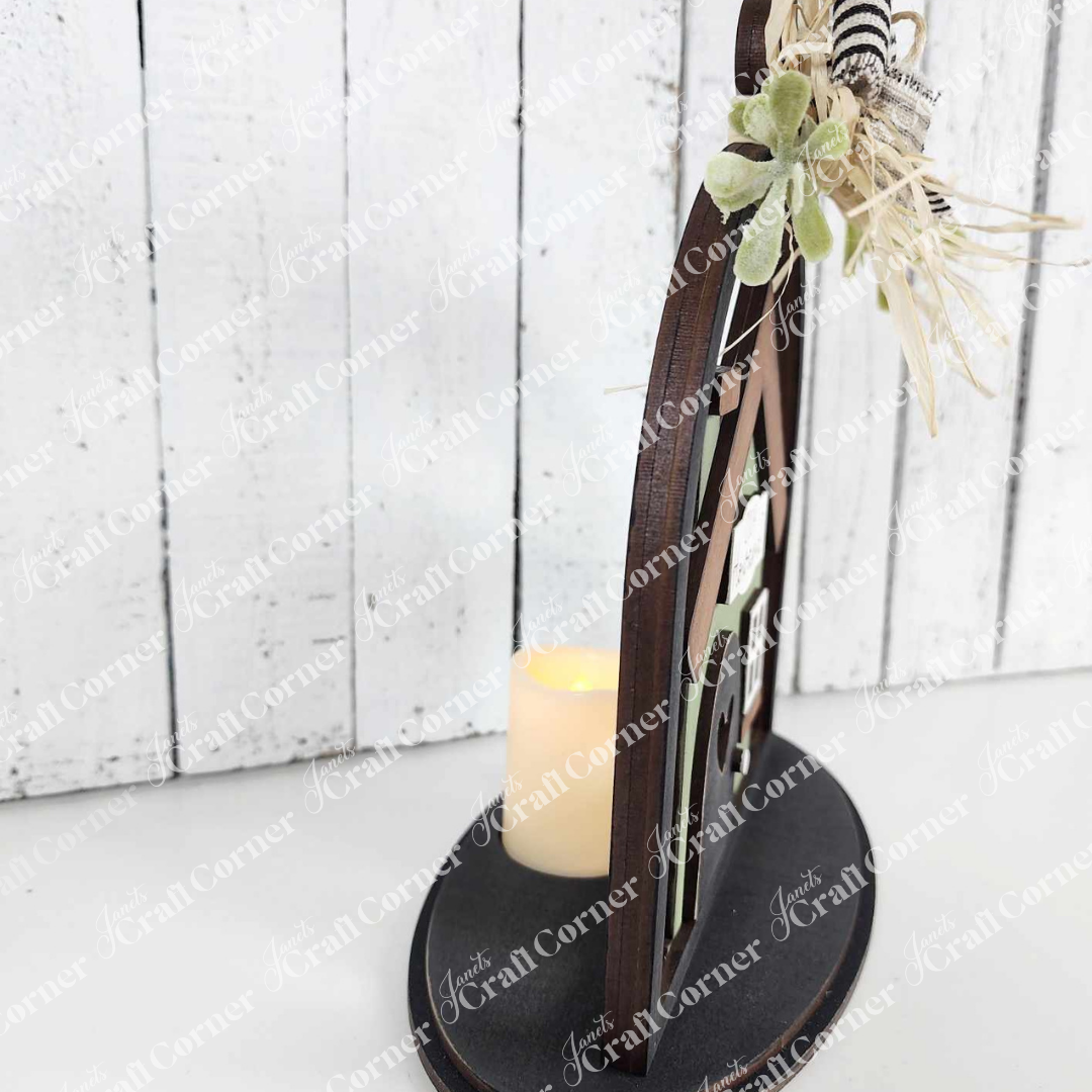 The Home Cloche Votive Holder Shelf Sitter from Janet's Craft Corner features a decorative wooden frame with green plants and a bow on an oval base, evoking a DIY craft kit. A battery-operated candle serves as a word art votive holder against a charming whitewashed wooden wall backdrop.