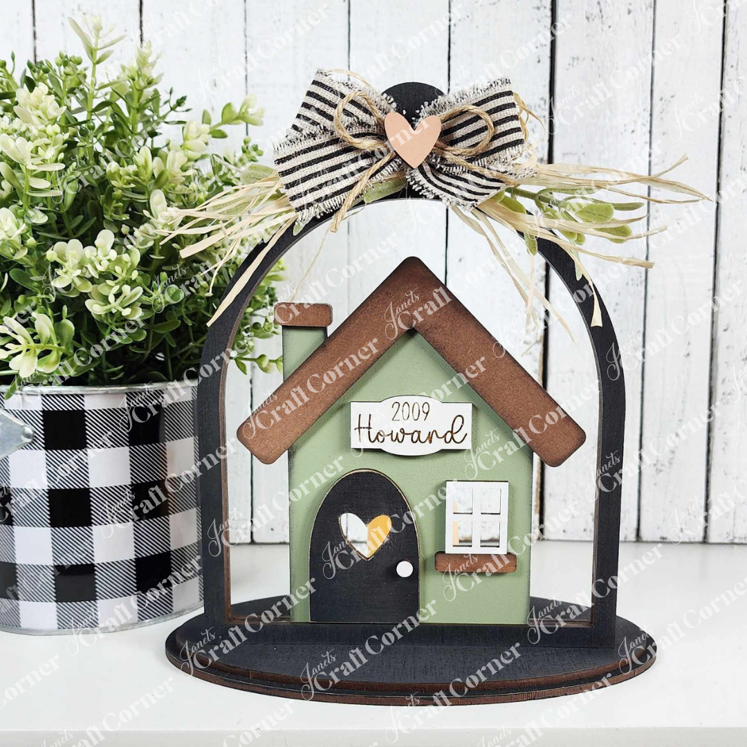 The "Home Cloche Votive Holder Shelf Sitter" by Janet's Craft Corner is a charming DIY home decor piece featuring a small house with "2009 Howard," adorned with a plaid bow, straw embellishment, and heart. Accompanied by a black and white checkered pot with greenery, it sits beautifully in any rustic display.
