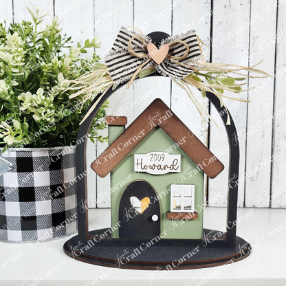 The "Home Cloche Votive Holder Shelf Sitter" by Janet's Craft Corner is a charming DIY home decor piece featuring a small house with "2009 Howard," adorned with a plaid bow, straw embellishment, and heart. Accompanied by a black and white checkered pot with greenery, it sits beautifully in any rustic display.