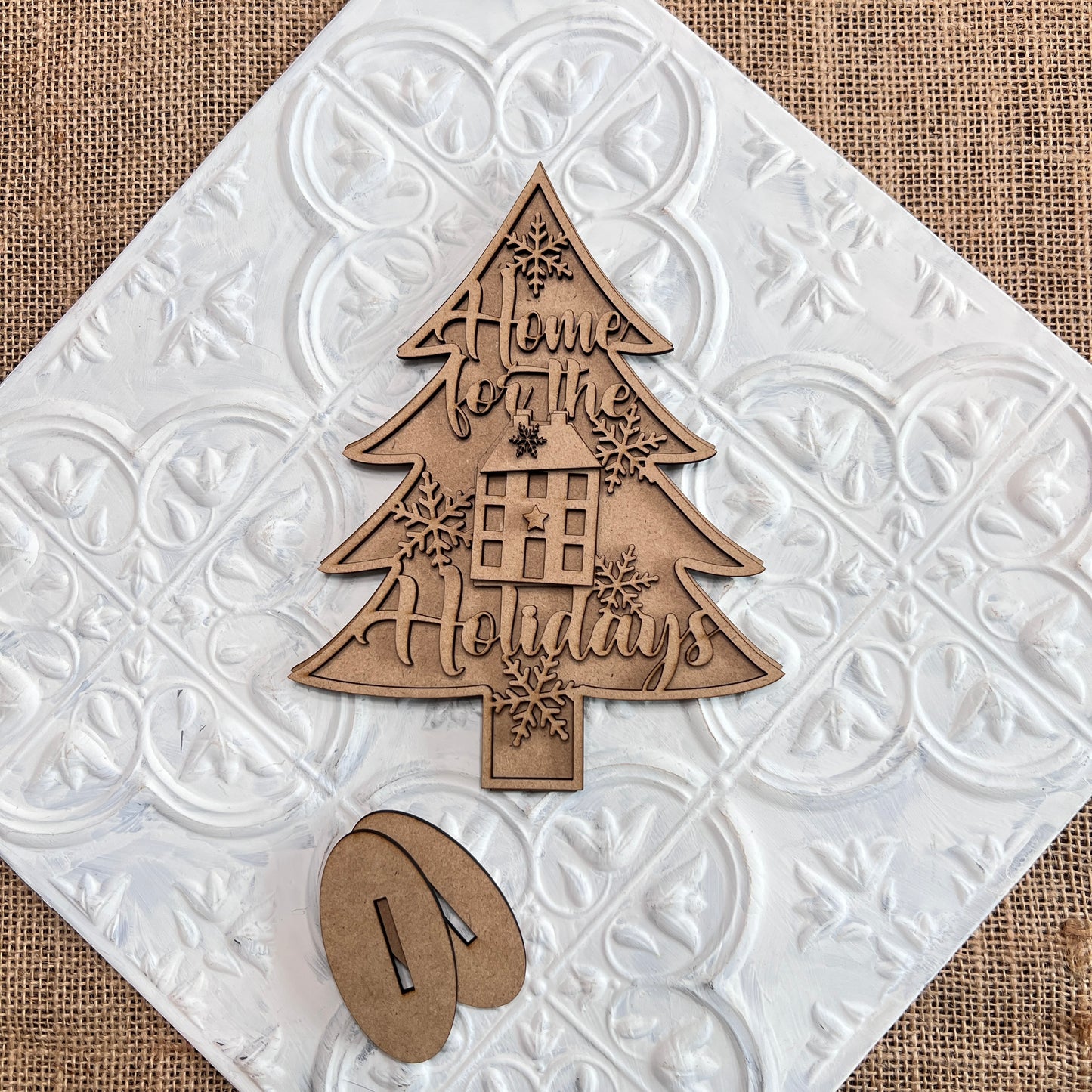 The Standing Christmas Tree Shelf Sitters from Janet's Craft Corner are perfect for your holiday decor. This wooden ornament, shaped like a Christmas tree with "Home for the Holidays" engraved on it, features snowflake designs and a window with a gift. It rests on a decorative white tile with burlap underneath—ideal for any shelf display.