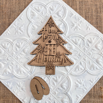 The Standing Christmas Tree Shelf Sitters from Janet's Craft Corner are perfect for your holiday decor. This wooden ornament, shaped like a Christmas tree with "Home for the Holidays" engraved on it, features snowflake designs and a window with a gift. It rests on a decorative white tile with burlap underneath—ideal for any shelf display.