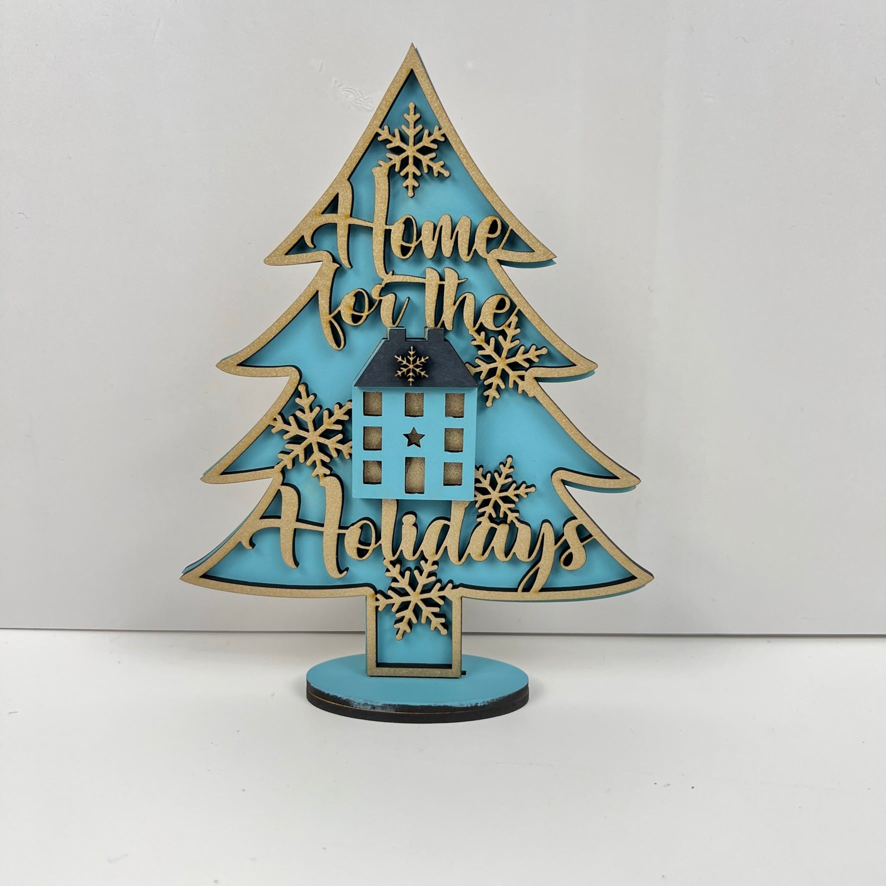 Introducing the "Standing Christmas Tree Shelf Sitters" by Janet's Craft Corner, a delightful piece of holiday decor. This wooden shelf display showcases a light blue tree shape adorned with intricate cut-outs of a house and snowflakes at its center, along with the phrase "Home for the Holidays," creating a charming festive atmosphere.