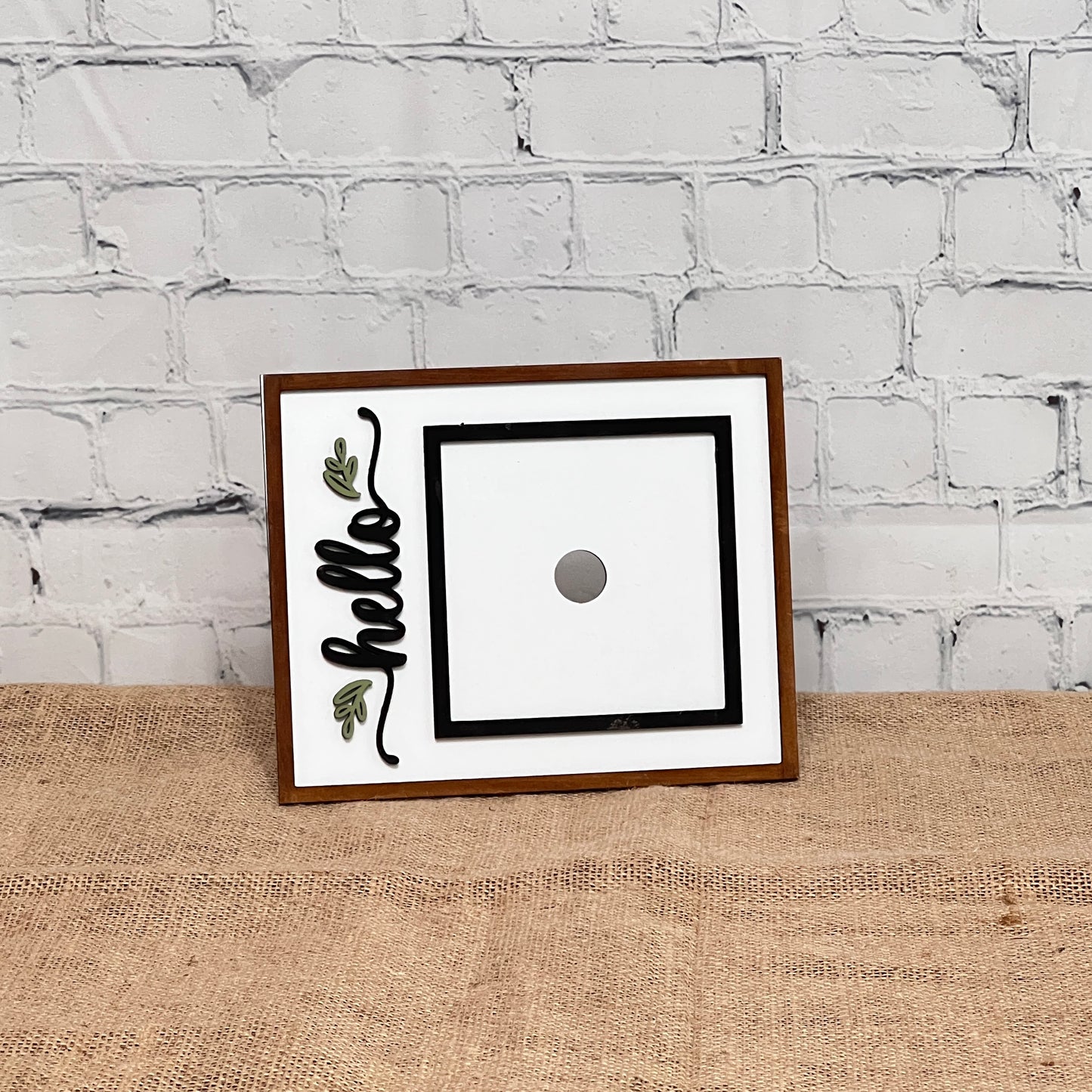 The Heartland Hello Swappable Squares Holder features a rectangular wooden frame with "hello" in black cursive, green leaf accents, and a center magnet for holding squares. It stands on burlap against a white brick wall.
