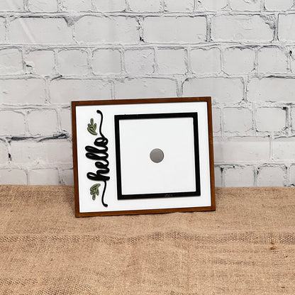 The Heartland Hello Swappable Squares Holder features a rectangular wooden frame with "hello" in black cursive, green leaf accents, and a center magnet for holding squares. It stands on burlap against a white brick wall.