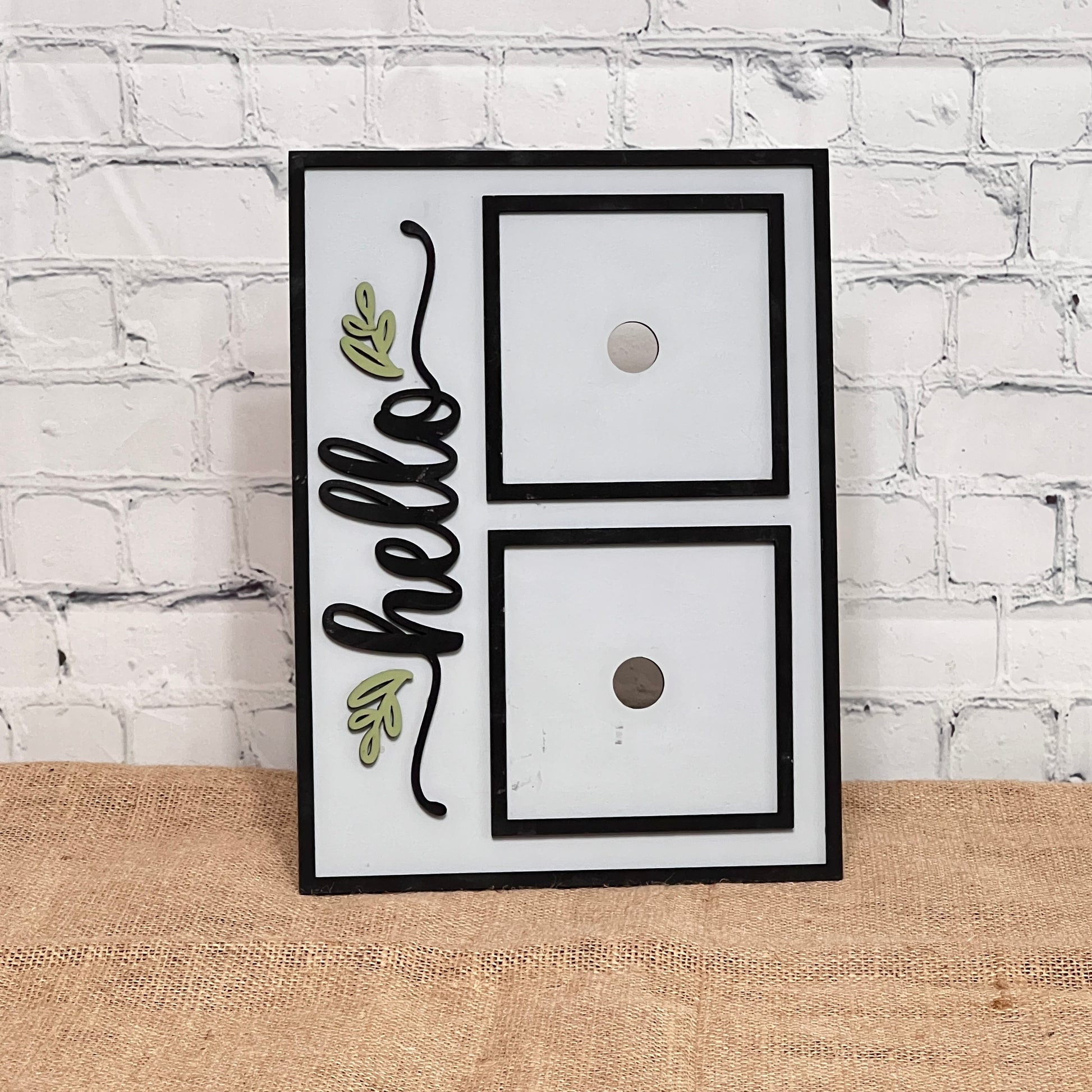 The Heartland Hello Swappable Squares Holder - Painted with Squares features "hello" in black cursive with green leaf accents on the left and two swappable square slots on the right, set against a white brick wall and burlap surface.