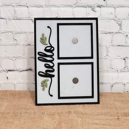 The Heartland Hello Swappable Squares Holder - Painted with Squares features "hello" in black cursive with green leaf accents on the left and two swappable square slots on the right, set against a white brick wall and burlap surface.