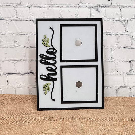 The Heartland Hello Swappable Squares Holder - Painted with Squares features "hello" in black cursive with green leaf accents on the left and two swappable square slots on the right, set against a white brick wall and burlap surface.