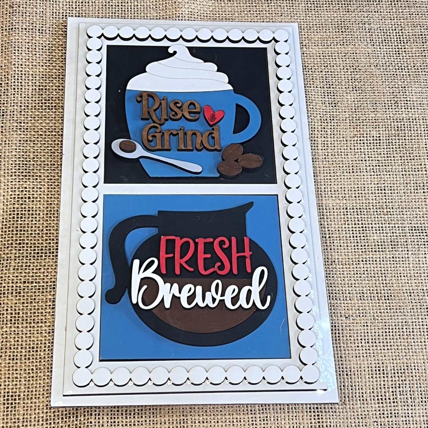 The Heartland Bead Trim Swappable Squares Frame with Inserts features two burlap panels: the top shows a mug with “Rise & Grind,” whipped cream, spoon, and beans; the bottom depicts a coffee pot with “Fresh Brewed.”.