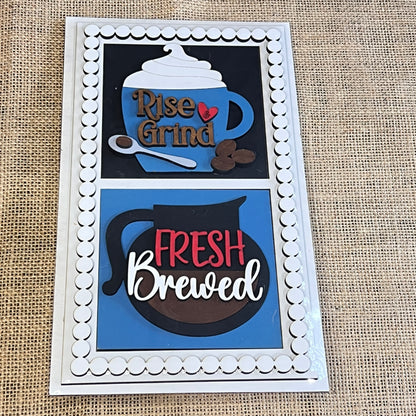 The Heartland Bead Trim Swappable Squares Frame with Inserts features two burlap panels: the top shows a mug with “Rise & Grind,” whipped cream, spoon, and beans; the bottom depicts a coffee pot with “Fresh Brewed.”.