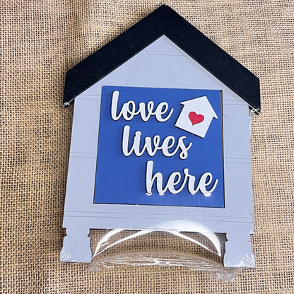 The Heartland House Swappable Squares Holders Shelf Leaner - Painted with Inserts features a house-shaped design on burlap with the phrase “love lives here” in white letters against a blue background, accented by a white arrow and red heart above “lives.” Crafted with wood cut-outs for added dimension, this charming shelf leaner blends rustic textures with heartfelt sentiment.