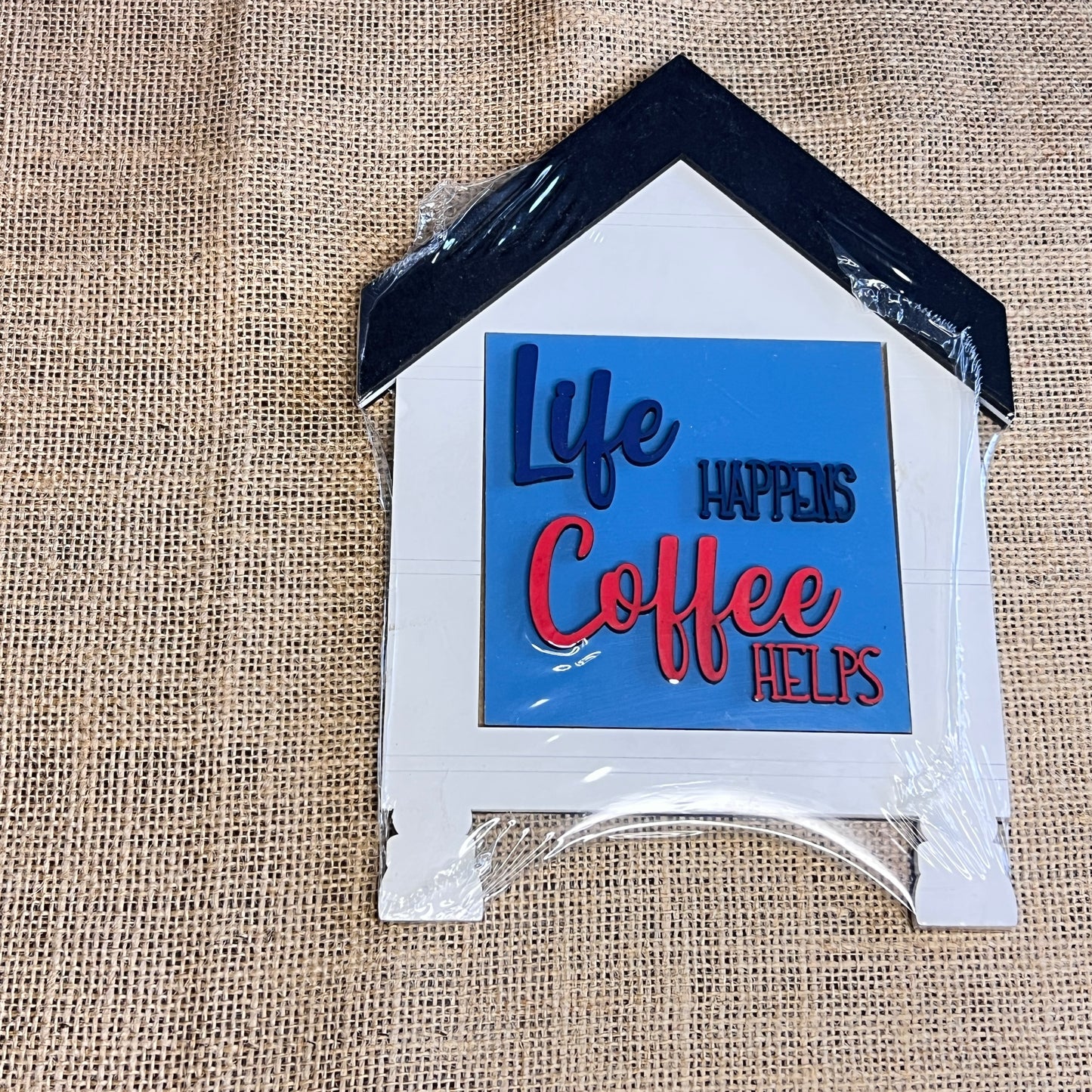 A Heartland House Swappable Squares Holders Shelf Leaner—painted with inserts—features a house-shaped design with “Life happens, coffee helps” in blue and red lettering on burlap, a black roof, and plastic-wrapped corners. Ideal for pairing with wood cut-outs or as part of a house frames base craft kit.