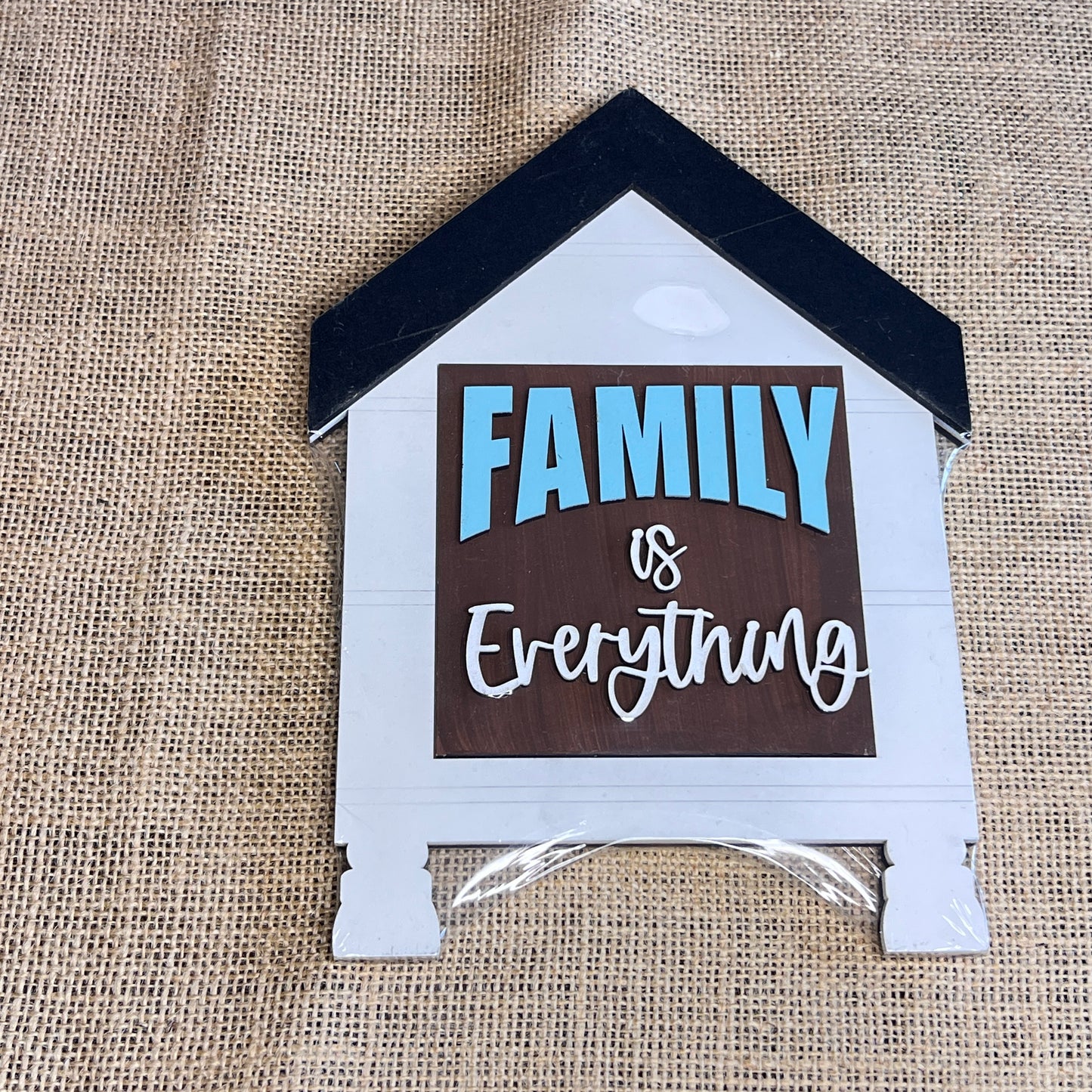 A Heartland House Swappable Squares Holders Shelf Leaner—painted in blue, white, and brown—features the phrase “FAMILY is Everything” on a burlap background. The roof is finished in black, and the bottom insert, crafted from one of our signature wood cut-outs, comes neatly wrapped in plastic.