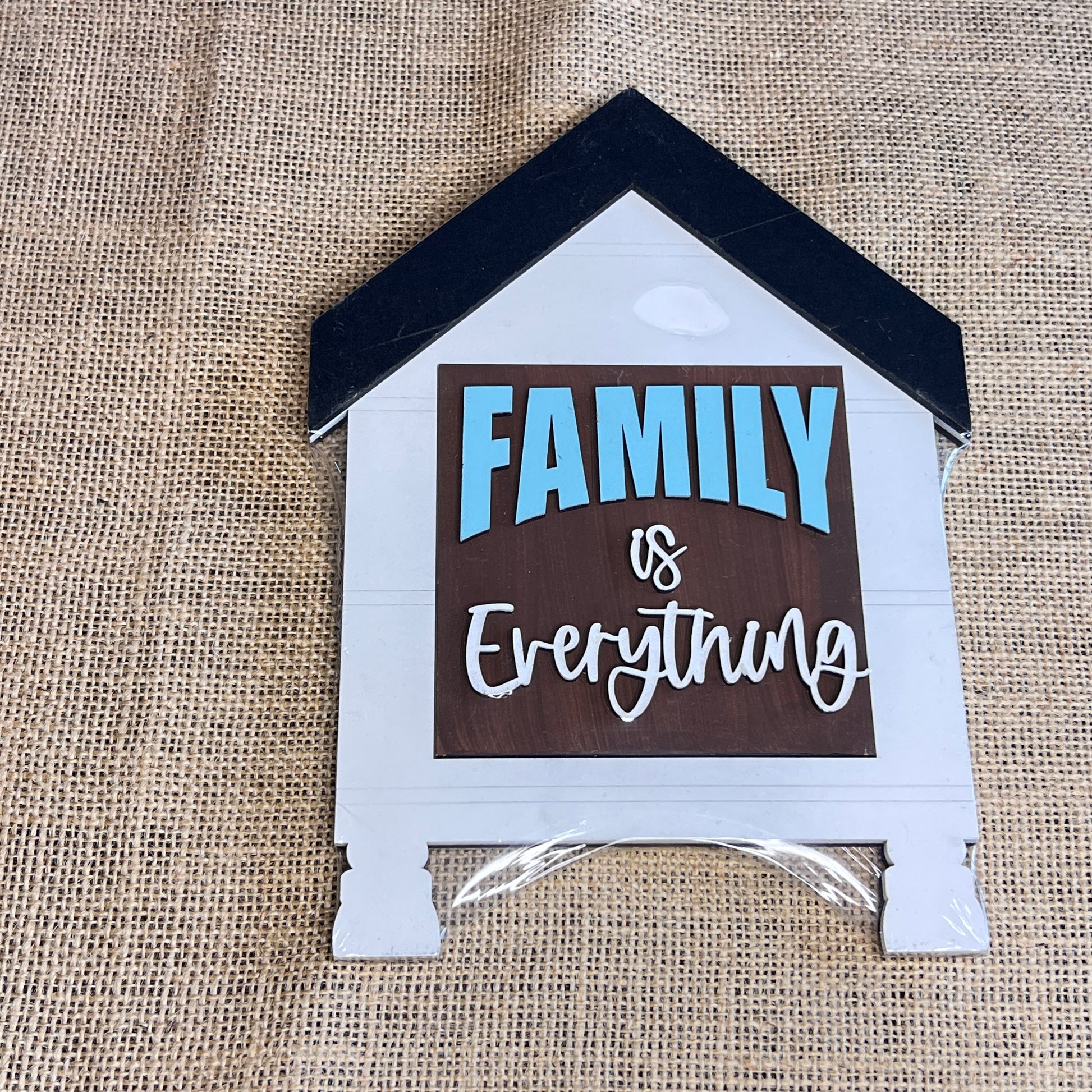 A Heartland House Swappable Squares Holders Shelf Leaner—painted in blue, white, and brown—features the phrase “FAMILY is Everything” on a burlap background. The roof is finished in black, and the bottom insert, crafted from one of our signature wood cut-outs, comes neatly wrapped in plastic.