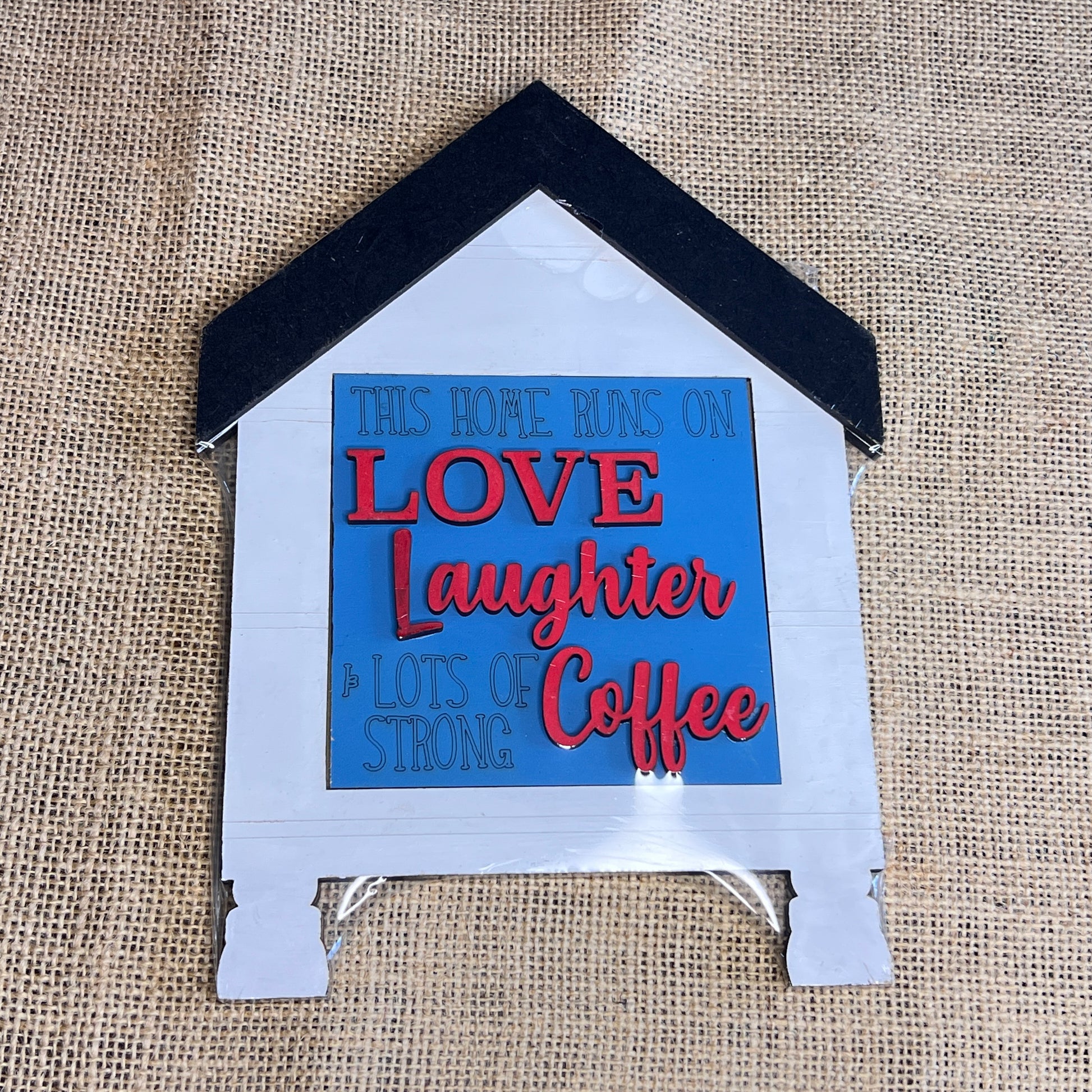 A delightful wooden “House Swappable Squares Holders Shelf Leaner - Painted with Inserts” by Heartland features a house-shaped design with the saying, “This home runs on LOVE, Laughter & lots of strong Coffee” on a blue background, displayed on a burlap surface.
