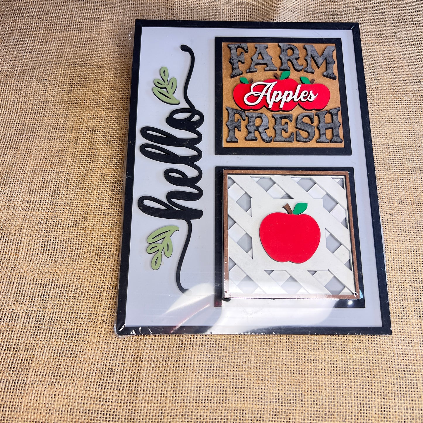 The Heartland Hello Swappable Squares Holder features three painted designs—a script "hello" with leaves, "FARM FRESH Apples," and a red apple—each as interchangeable wood inserts or charming shelf sitters.