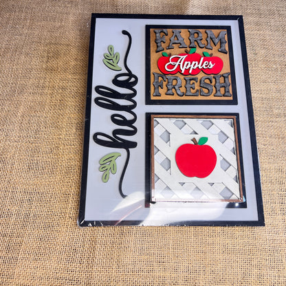 The Heartland Hello Swappable Squares Holder features three painted designs—a script "hello" with leaves, "FARM FRESH Apples," and a red apple—each as interchangeable wood inserts or charming shelf sitters.