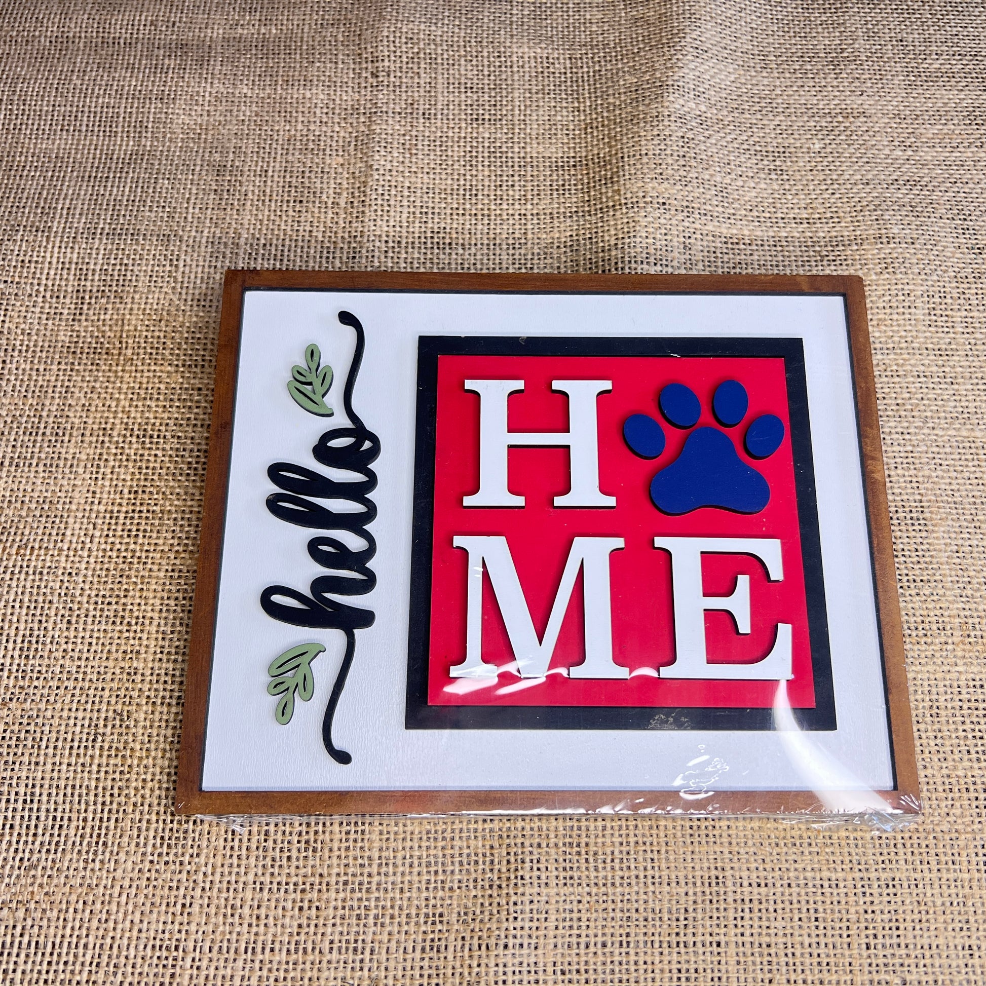 The Heartland Hello Swappable Squares Holder - Painted with Squares features "HOME" with a blue paw print "O" and "hello" with leafy accents on white burlap in a wooden frame—ideal as shelf décor or paired with wood square inserts.