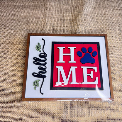 The Heartland Hello Swappable Squares Holder - Painted with Squares features "HOME" with a blue paw print "O" and "hello" with leafy accents on white burlap in a wooden frame—ideal as shelf décor or paired with wood square inserts.