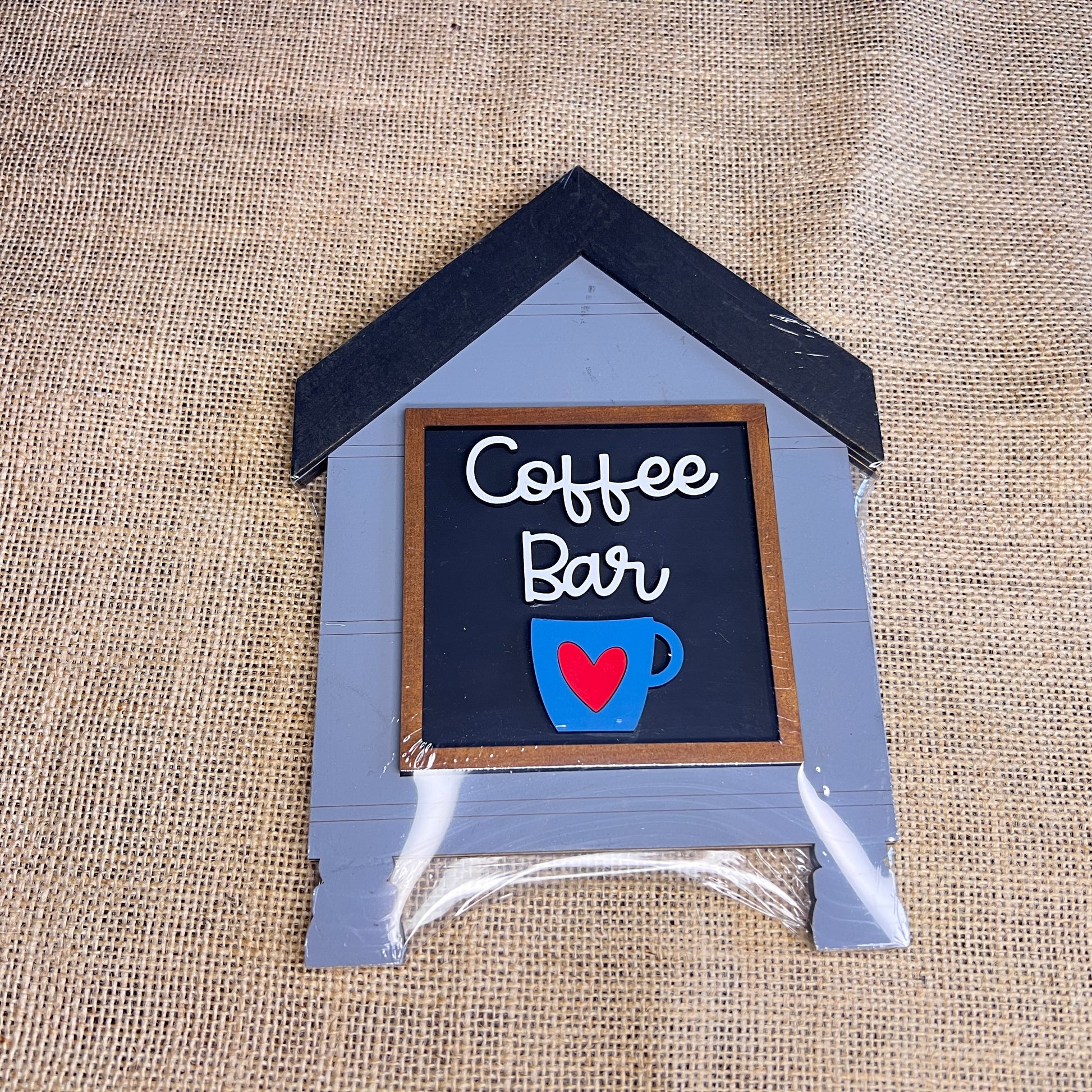 A small, house-shaped “House Swappable Squares Holders Shelf Leaner - Painted with Inserts” by Heartland, crafted from wood cut-outs on burlap, features “Coffee Bar” in white lettering above a blue coffee cup and red heart illustration.