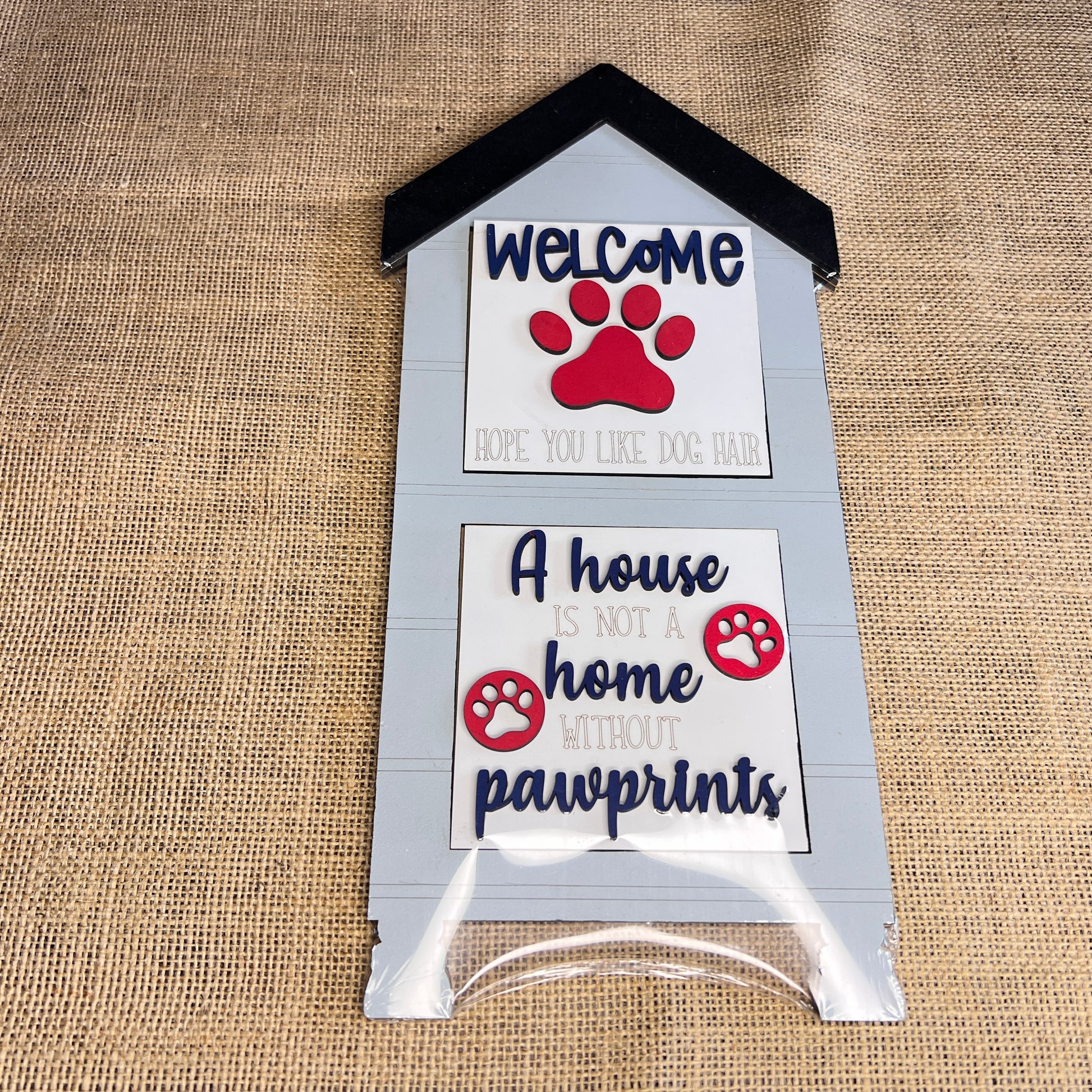 The Heartland House Swappable Squares Holders Shelf Leaner – Painted with Inserts features a doghouse-shaped wood cut-out sign with the message “Welcome, hope you like dog hair. A house is not a home without pawprints,” accented by red paw prints and displayed against a charming burlap background.