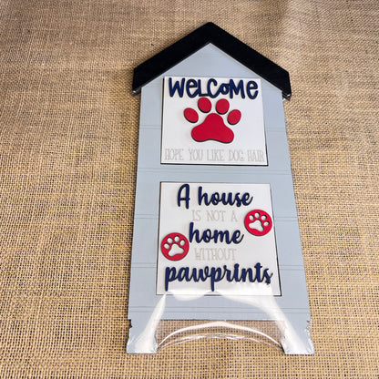 The Heartland House Swappable Squares Holders Shelf Leaner – Painted with Inserts features a doghouse-shaped wood cut-out sign with the message “Welcome, hope you like dog hair. A house is not a home without pawprints,” accented by red paw prints and displayed against a charming burlap background.