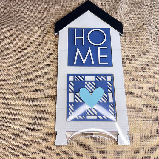 The Heartland House Swappable Squares Holders Shelf Leaner - Painted with Inserts is a house-shaped burlap sign with "HOME" in blue and white above a blue heart, perfect for DIY displays or use with wood square inserts.