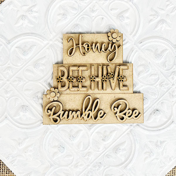 Cursive text on stacked wooden plaques spells out "Honey," "Bee Hive," and "Bumble Bee" against a textured white backdrop, exuding farmhouse-style charm. Small floral accents grace the top and bottom plaques, creating a delightful addition to your Summer Word Stacks collection by Janet's Craft Corner.