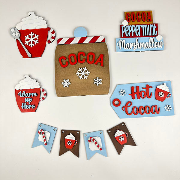 Experience a delightful holiday décor collection by Janet's Craft Corner, featuring cocoa-themed items. The Hot Cocoa Tiered Tray Pieces include red and blue signs with cozy phrases like "Warm up here," a charming brown bag labeled "COCOA," and images of hot cocoa cups topped with whipped cream and snowflakes.