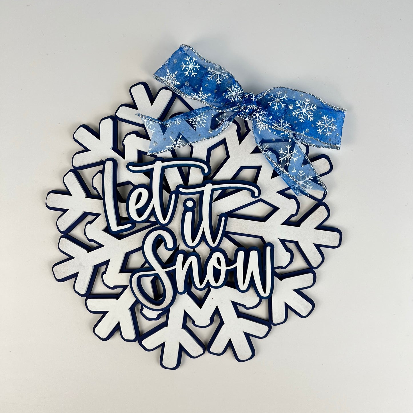 Introducing the Let it Snow Snowflake Door Hanger from Janet's Craft Corner: This decorative piece features bold "Let it Snow" lettering on a white snowflake design. Enhanced with a blue ribbon adorned with snowflake patterns, it's an ideal addition to seasonal décor, beautifully standing out against a neutral white background.