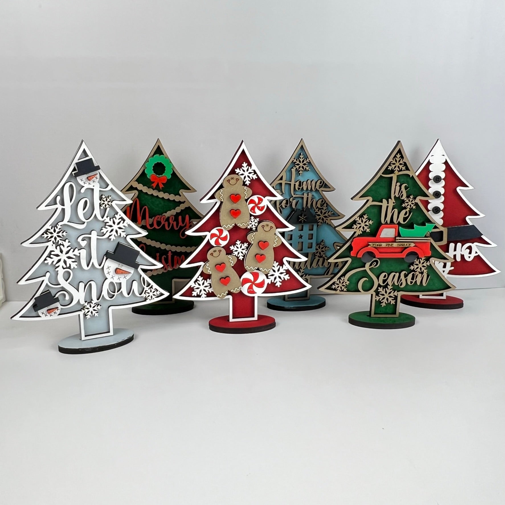 Introducing the Standing Christmas Tree Shelf Sitters by Janet's Craft Corner. These delightful holiday decorations come adorned with a variety of festive designs and messages such as "Let it Snow" and "Merry Christmas." Featuring motifs like snowflakes, candy canes, and a red truck, these charming pieces are perfect for enhancing your shelf display against a white background.