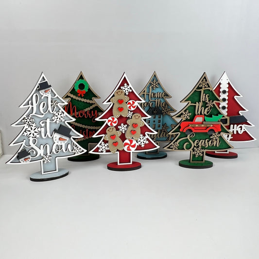 Introducing the Standing Christmas Tree Shelf Sitters by Janet's Craft Corner. These delightful holiday decorations come adorned with a variety of festive designs and messages such as "Let it Snow" and "Merry Christmas." Featuring motifs like snowflakes, candy canes, and a red truck, these charming pieces are perfect for enhancing your shelf display against a white background.