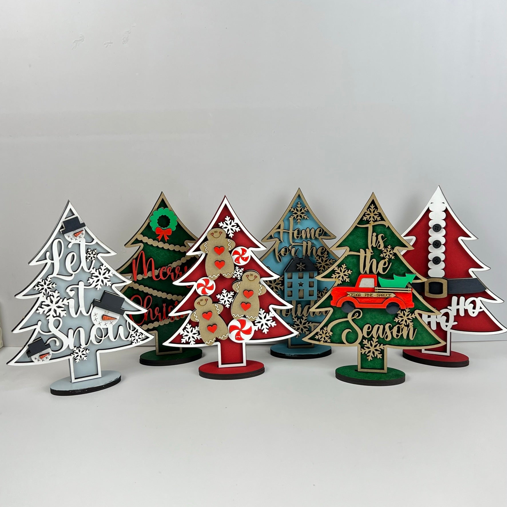 Explore the Standing Christmas Tree Shelf Sitters by Janet's Craft Corner, a collection featuring festive designs and messages such as "Let it Snow" and "Home for the Holidays." These vibrant wooden trees bring cheer to any shelf display with delightful details like snowflakes, wreaths, and candy canes enhancing your holiday decor.