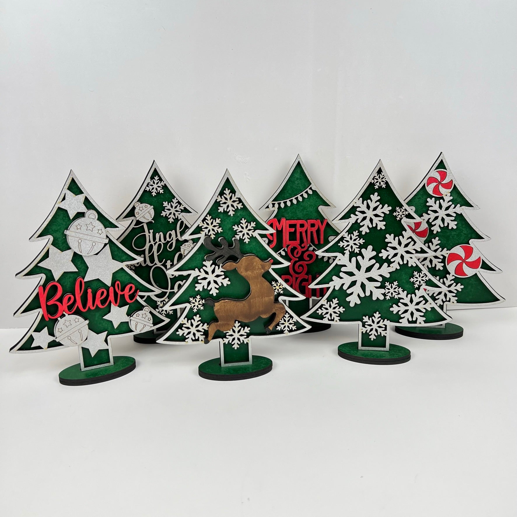 Introducing Janet's Craft Corner's Standing Christmas Tree Shelf Sitters, a charming collection of decorative wooden Christmas trees. Featuring the words "Believe," "Jingle," and "Merry," these sitters boast delightful embellishments such as snowflakes, ornaments, a reindeer, and candy canes on a vibrant green background. They are perfect for enhancing your holiday decor and would make an ideal addition to any festive shelf display.