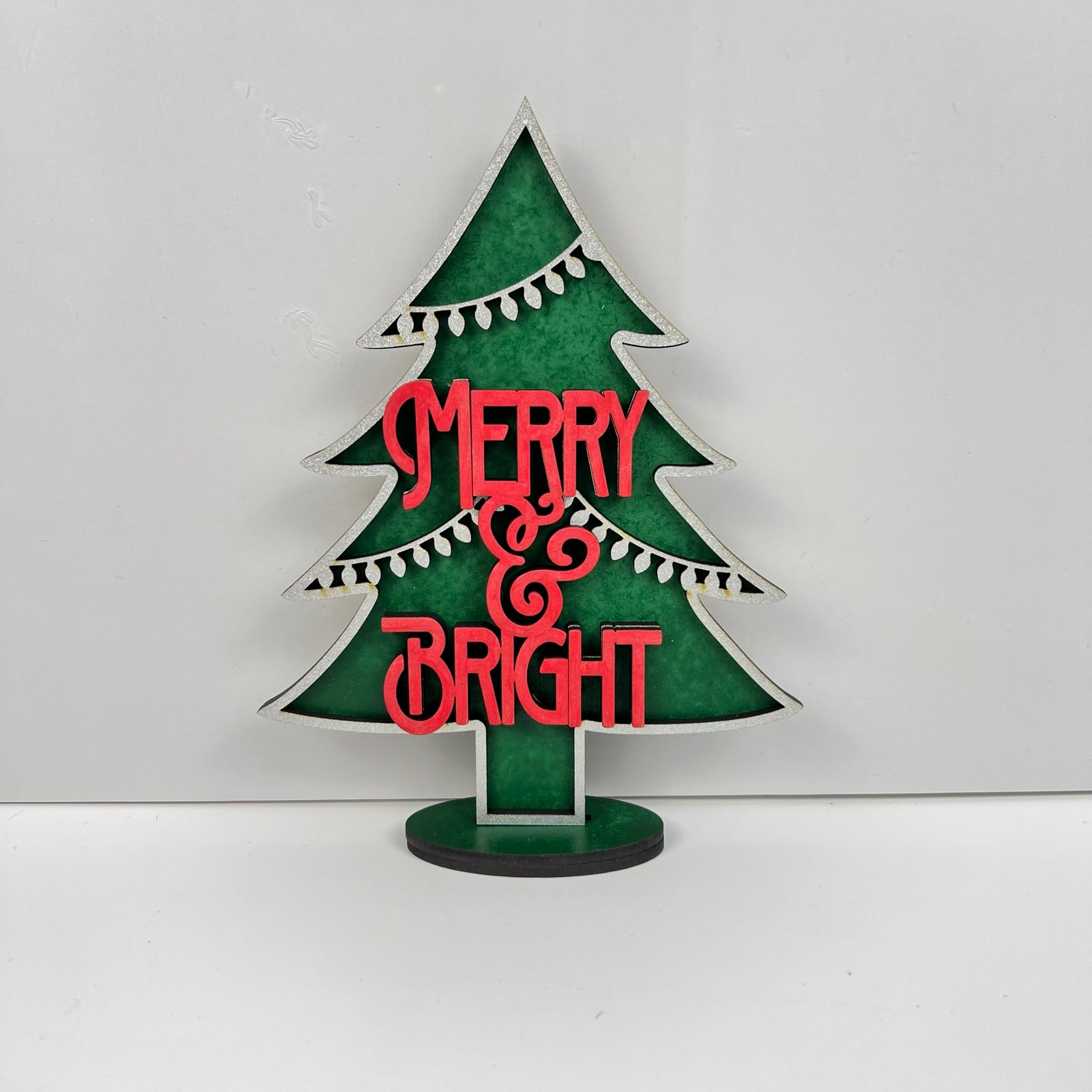 The "Standing Christmas Tree Shelf Sitters" by Janet's Craft Corner is a delightful decorative piece perfect for shelf displays. It features a green background with the phrase "Merry & Bright" in red, and its edges are adorned with white bulb designs that resemble string lights. This charming item brings festive flair to any holiday decor setting.