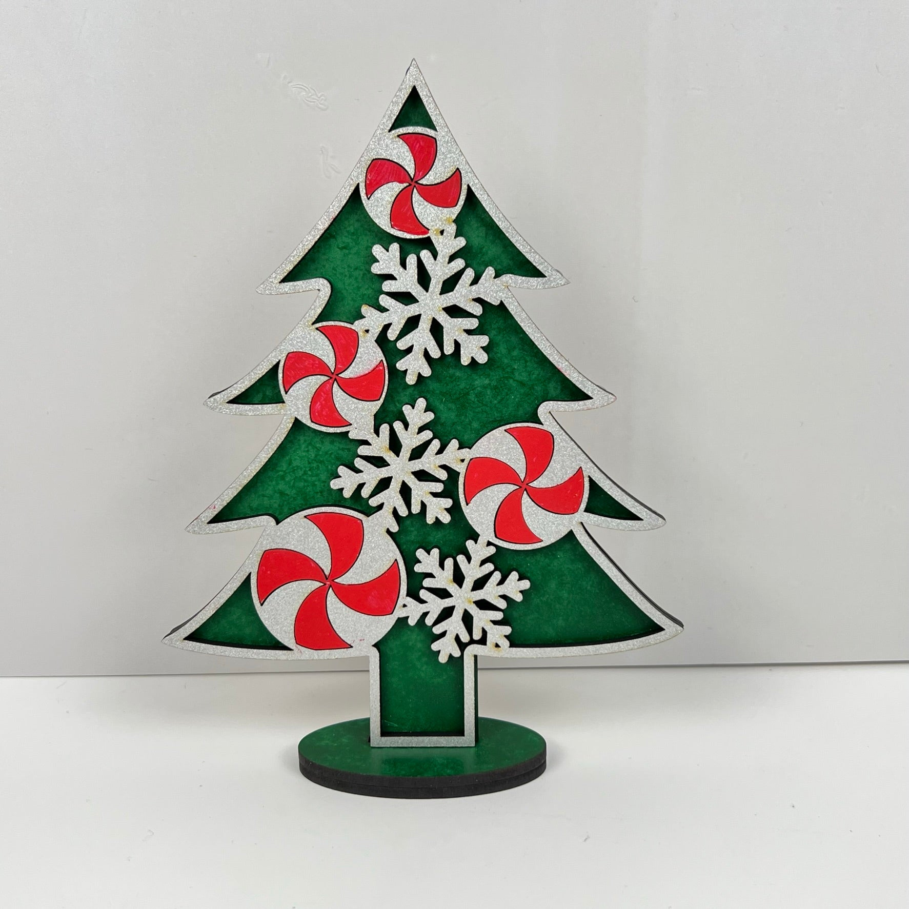 Introducing Janet's Craft Corner's Standing Christmas Tree Shelf Sitters - a perfect addition to your holiday decor. This piece features a green Christmas tree cutout, elegantly decorated with red and white swirl ornaments reminiscent of peppermint candies and delicate white snowflakes. Designed for shelf display, it stands securely on a round base against any plain background, making it an ideal festive accent for your home.