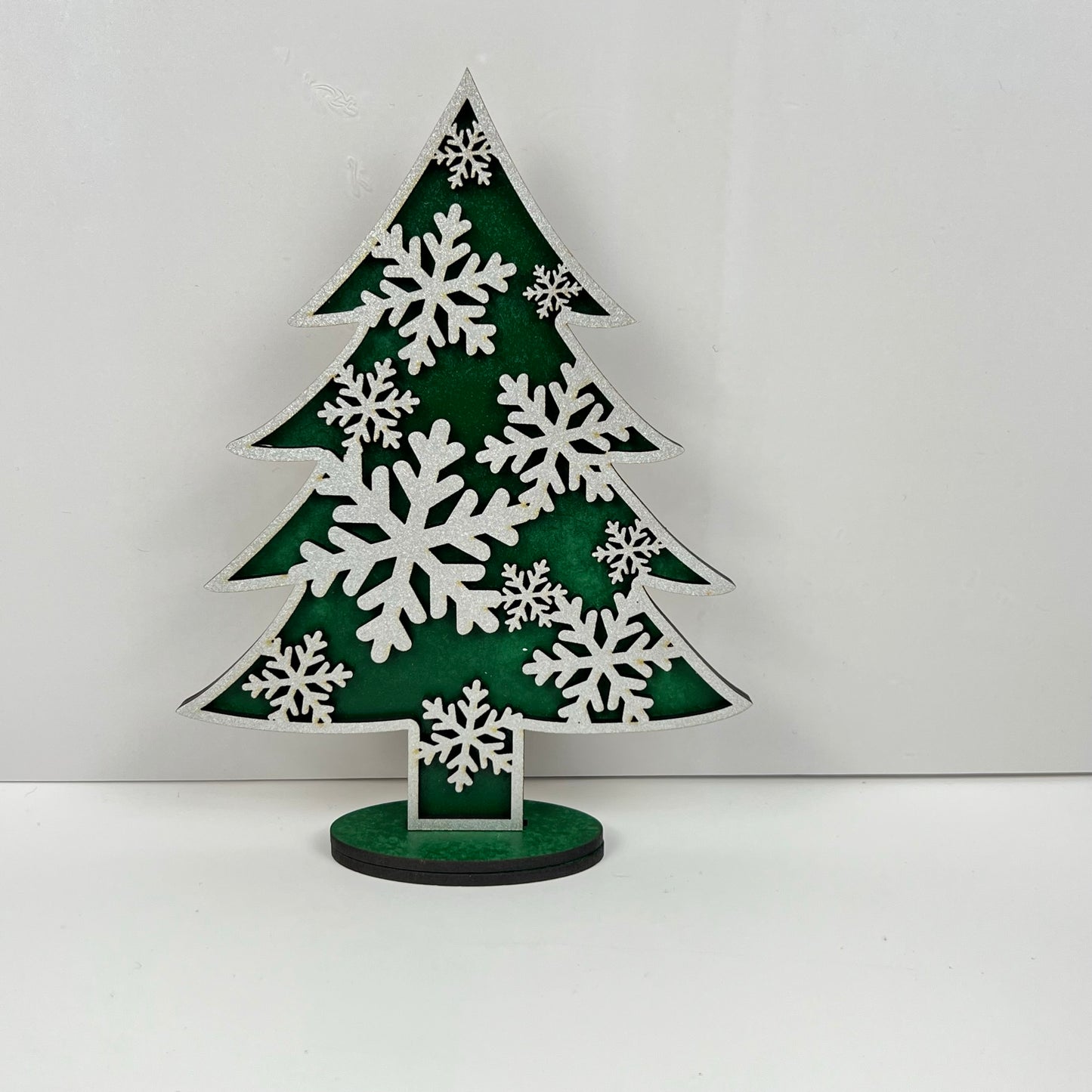 The "Standing Christmas Tree Shelf Sitters" from Janet's Craft Corner make for a perfect holiday decor piece. This decorative green ornament is shaped like a Christmas tree and adorned with white snowflakes, standing out beautifully against a plain white background. It's ideal for adding an eye-catching touch to your festive shelf display.