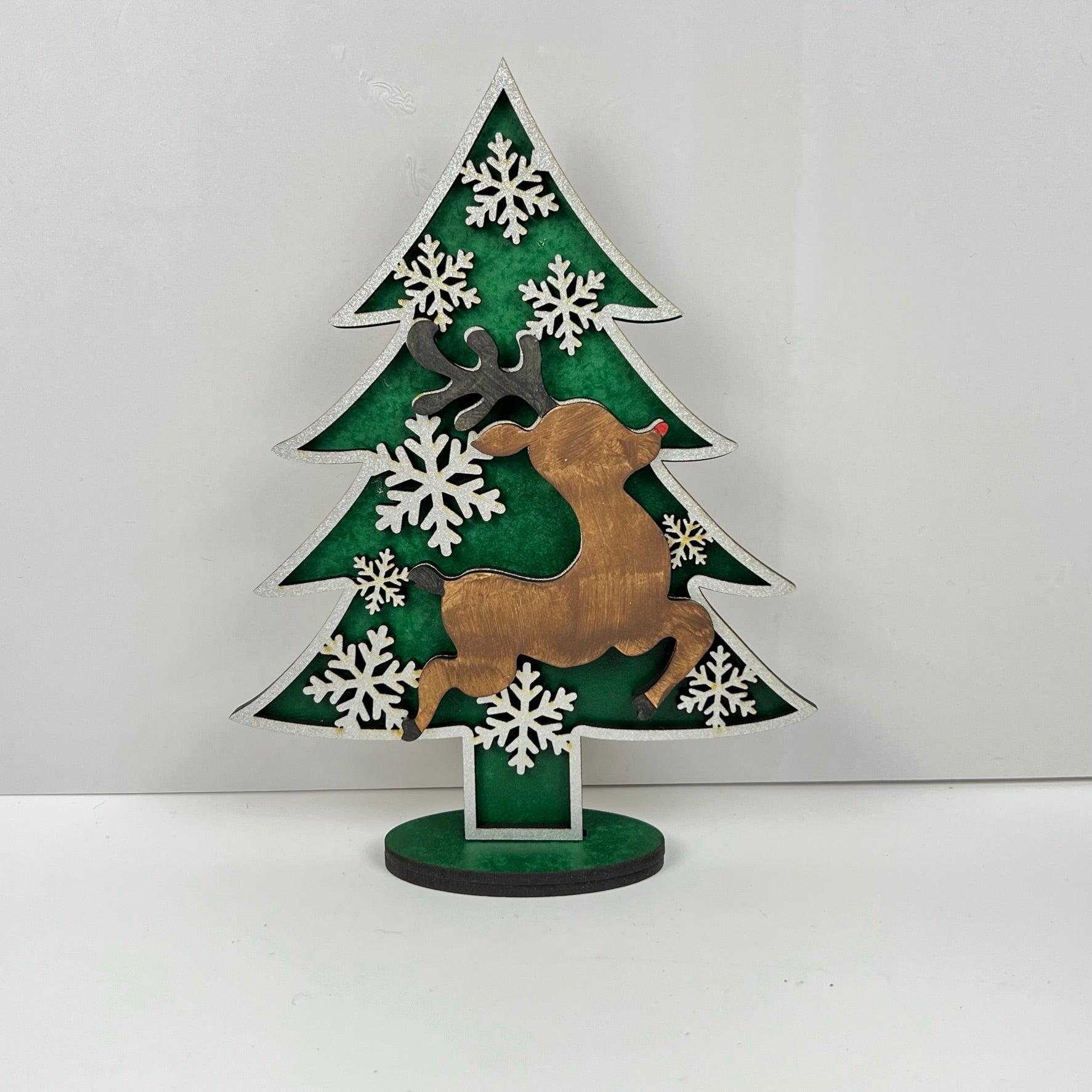 This holiday decor piece is a Standing Christmas Tree Shelf Sitter from Janet's Craft Corner, featuring a green background adorned with a brown reindeer silhouette and white snowflakes. It's perfect for adding festive charm to your shelf display, standing proudly on its circular base.