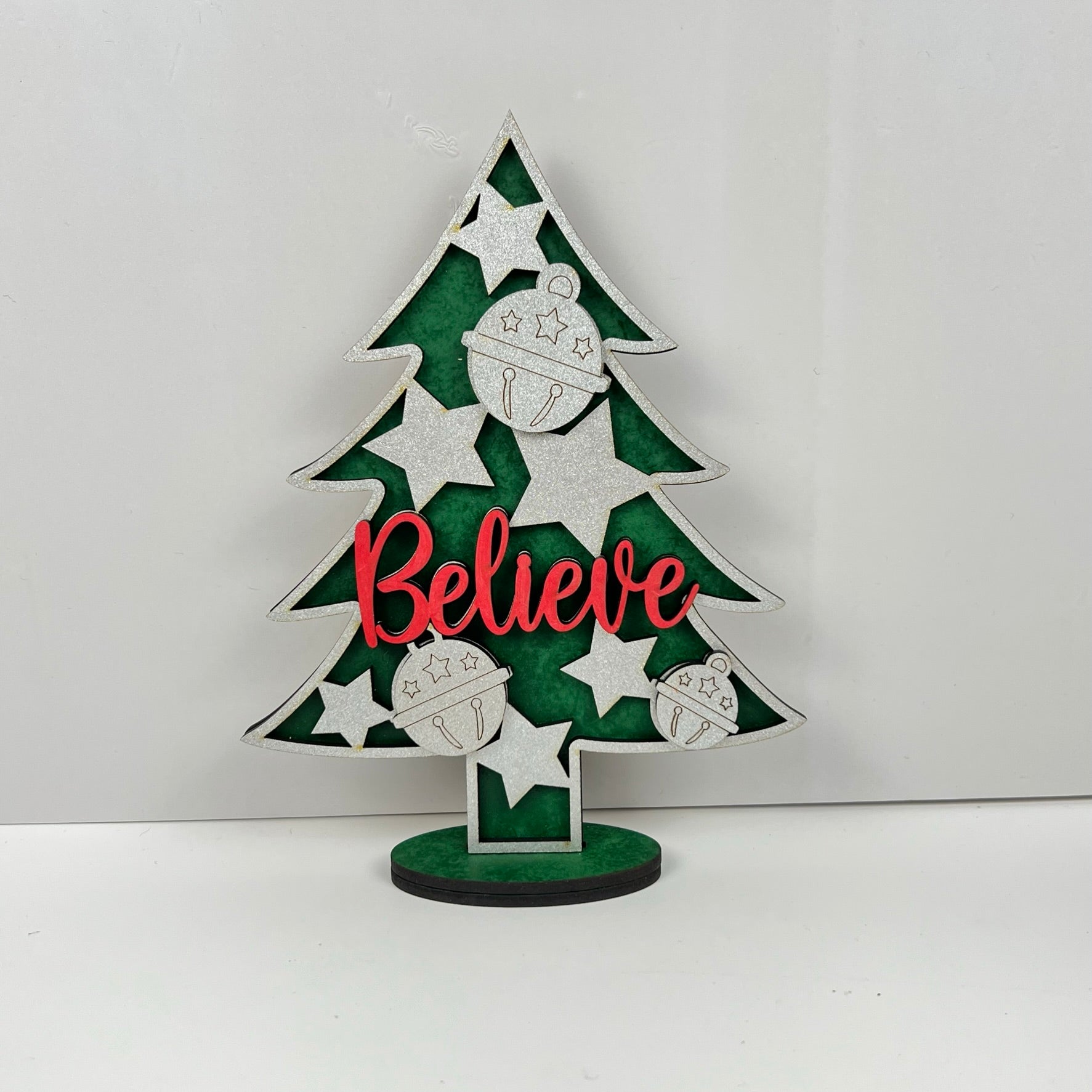 Introducing the Standing Christmas Tree Shelf Sitters by Janet's Craft Corner. This festive decoration features a beautifully designed Christmas tree cutout adorned with silver stars and ornaments. Highlighted with the word "Believe" in bold red across the center, it adds a charming holiday touch to your decor, especially against a plain white wall. Perfect for enhancing any shelf display this holiday season.