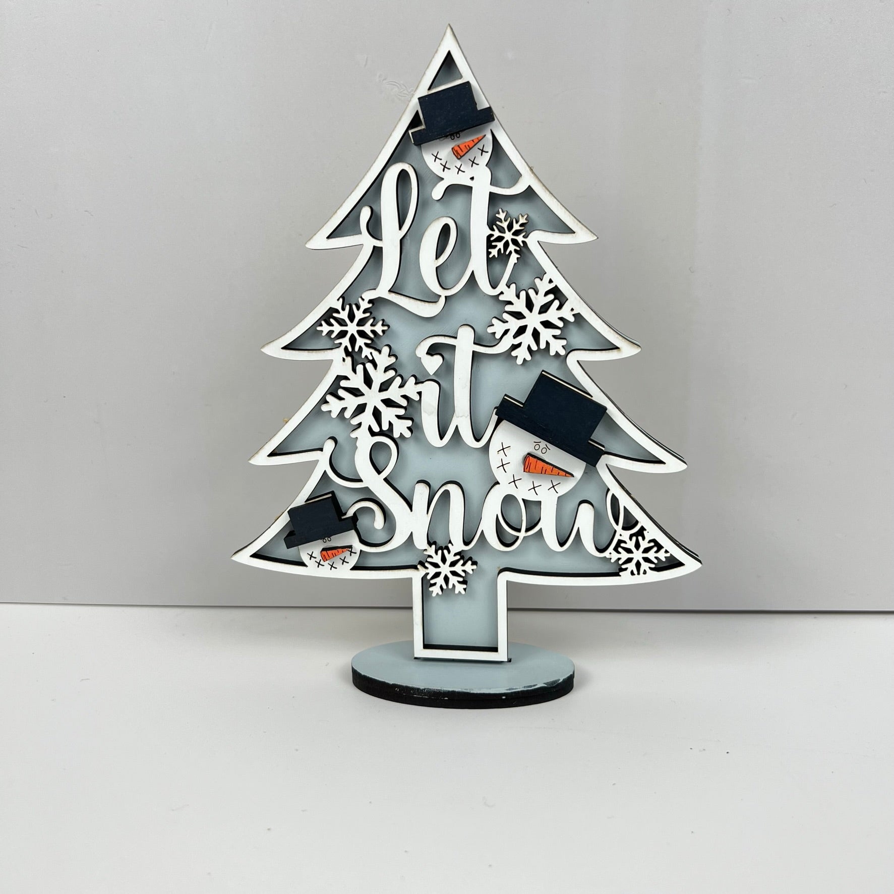 Introducing the Standing Christmas Tree Shelf Sitters by Janet's Craft Corner—a delightful addition to your holiday decor. This decorative piece features the phrase "Let it Snow" in elegant cursive, adorned with three small snowmen in black hats and snowflake accents on a light blue background, making it perfect for your Christmas tree or as a charming shelf display.