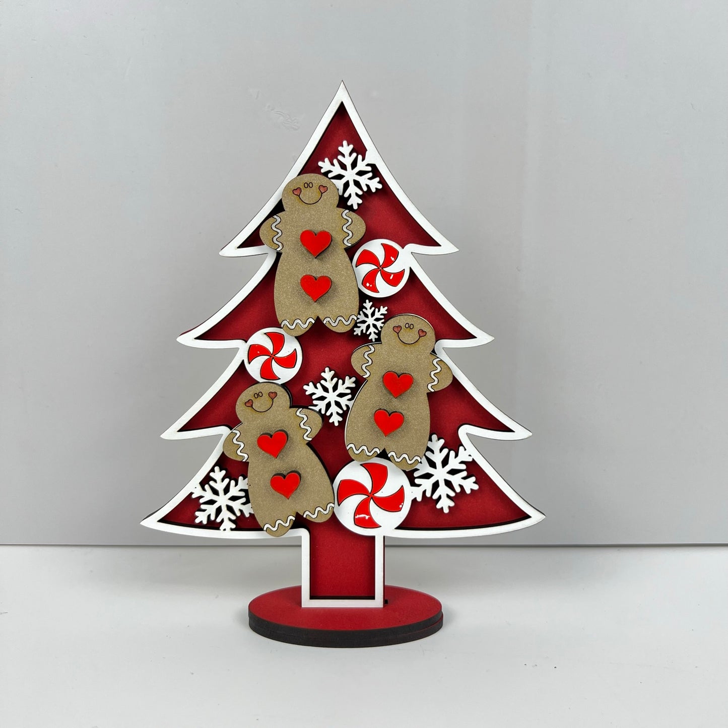 Introducing the "Standing Christmas Tree Shelf Sitters" from Janet's Craft Corner. This charming holiday decor features gingerbread men, red and white peppermint wheels, and white snowflakes atop a festive red background with a white outline. Ideal for any seasonal shelf display, it stands gracefully on a small circular base.