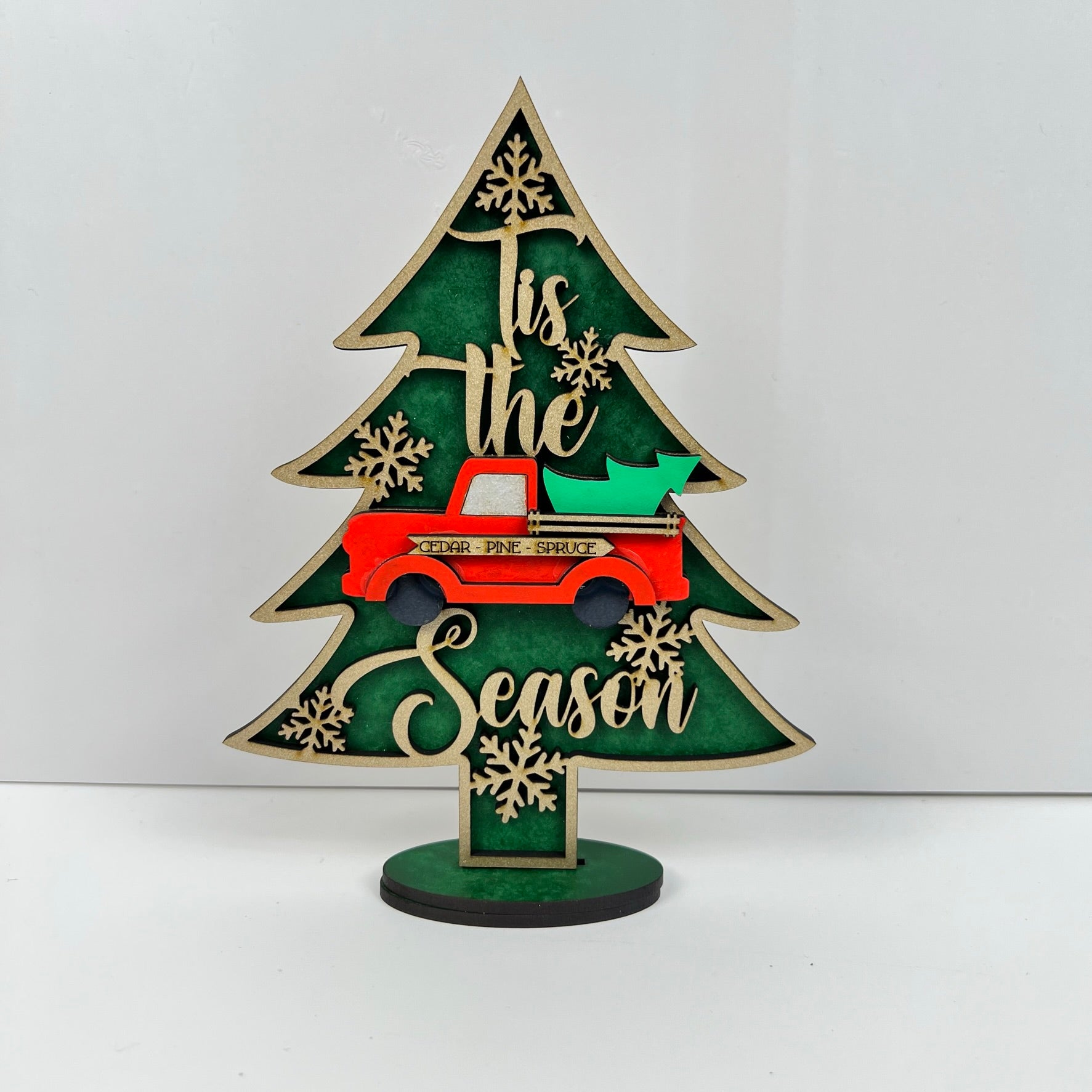 The Standing Christmas Tree Shelf Sitters by Janet's Craft Corner is a decorative wooden piece that features the phrase "Tis the Season" and showcases a red truck with a green tree at its center. It is adorned with snowflake cutouts against a green backdrop, making it perfect for your holiday decor and festive shelf display.