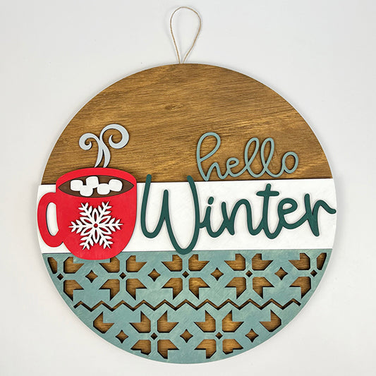 The Hello Winter Hot Cocoa Door Hanger by Janet's Craft Corner features a round wooden design with "Hello Winter" in teal script, adorned with a red mug of hot cocoa topped with marshmallows and a snowflake motif. The bottom half displays a blue geometric pattern, and it includes an attached string for easy hanging.