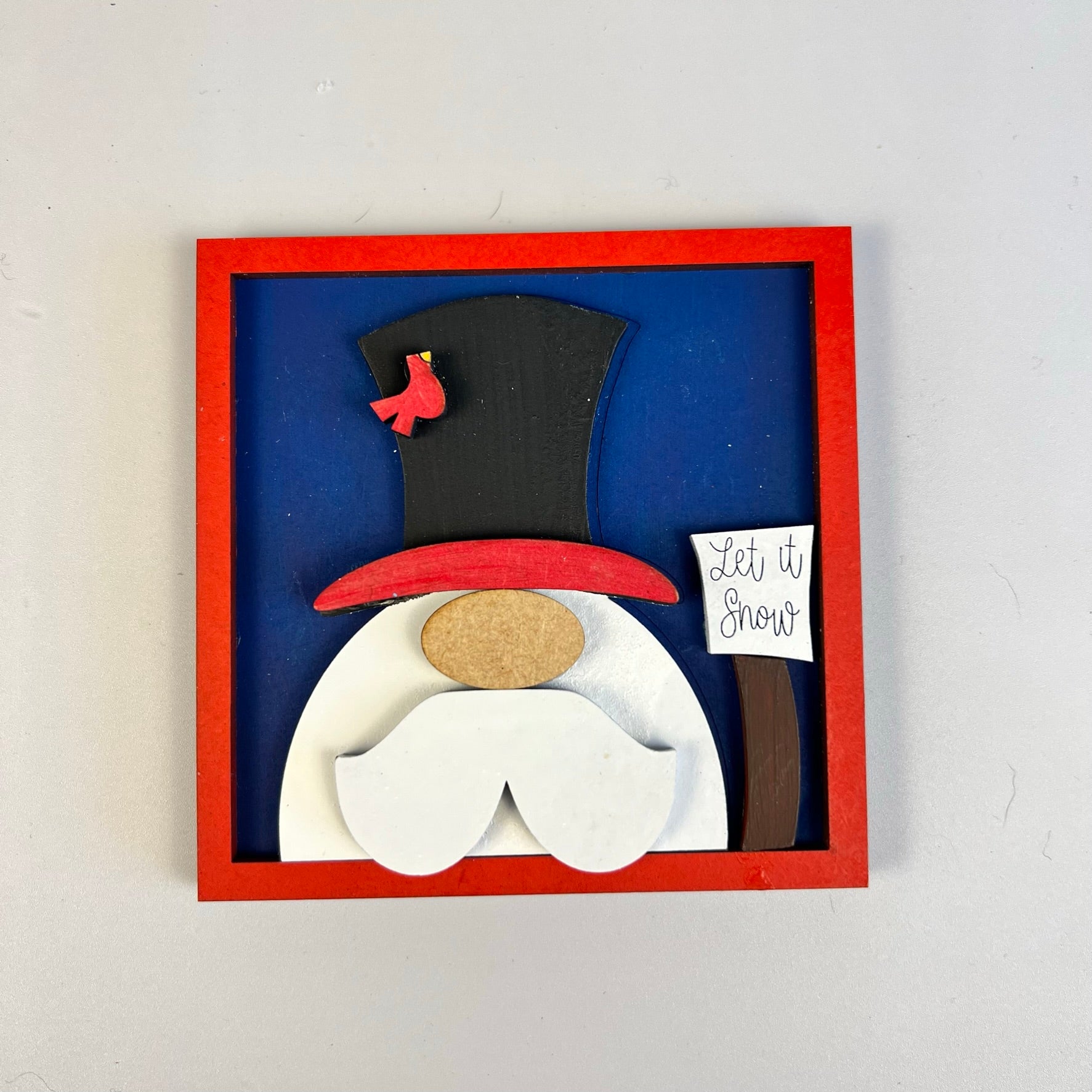 The Winter Interchangeable Squares from Janet's Craft Corner feature a charming festive gnome decoration inside a red frame, ideal for tiered trays. The gnome has a large white beard and wears a tall black hat topped with a small red bird. It holds a "Let it Snow" sign against the blue background, perfectly enhancing your seasonal décor.