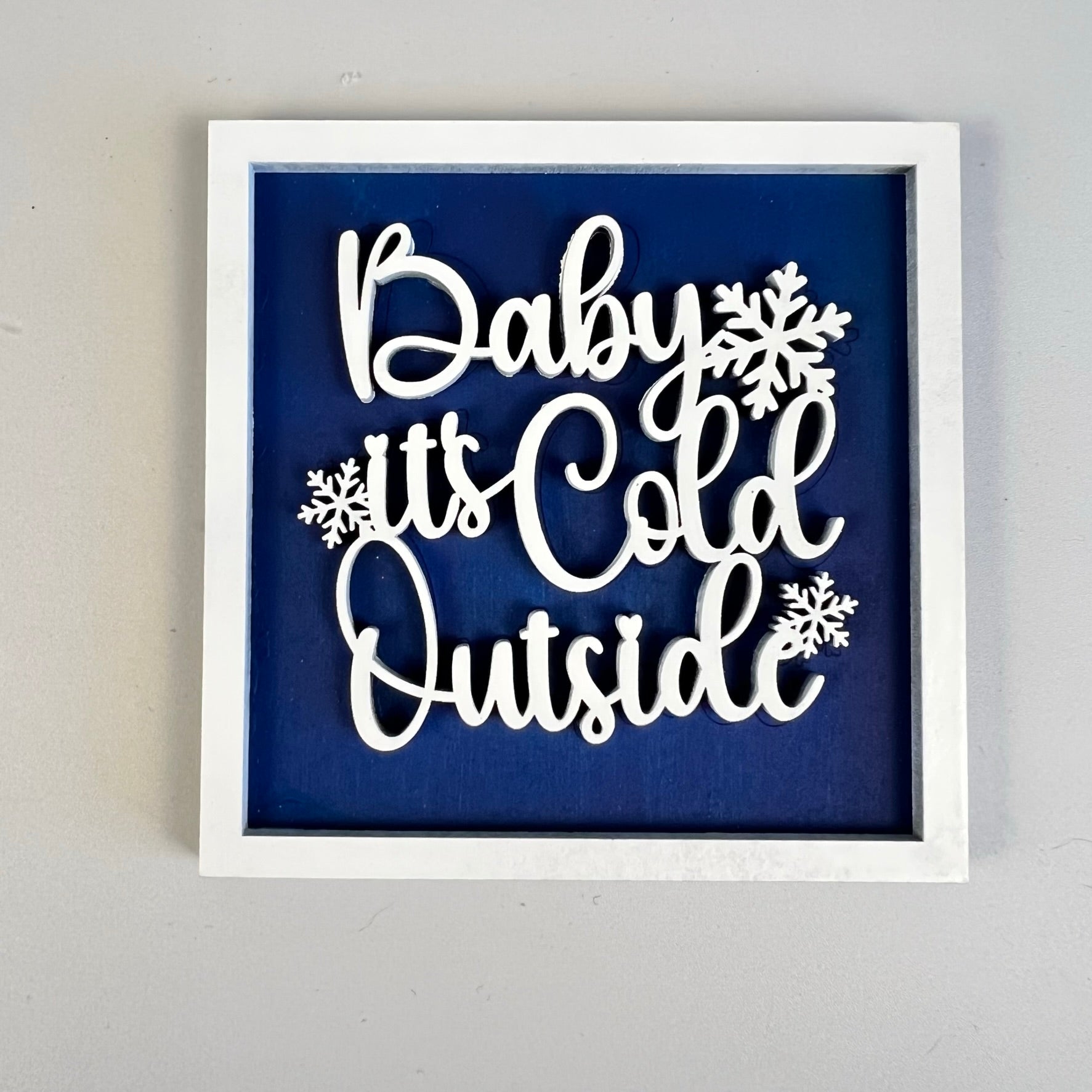 A framed sign from Janet's Craft Corner, featuring the "Winter Interchangeable Squares" design, displays white cursive text on a blue background that reads "Baby it's Cold Outside," adorned with snowflakes. Its white frame complements its seasonal décor style, making it an ideal addition to tiered trays and enhancing the wintry theme.