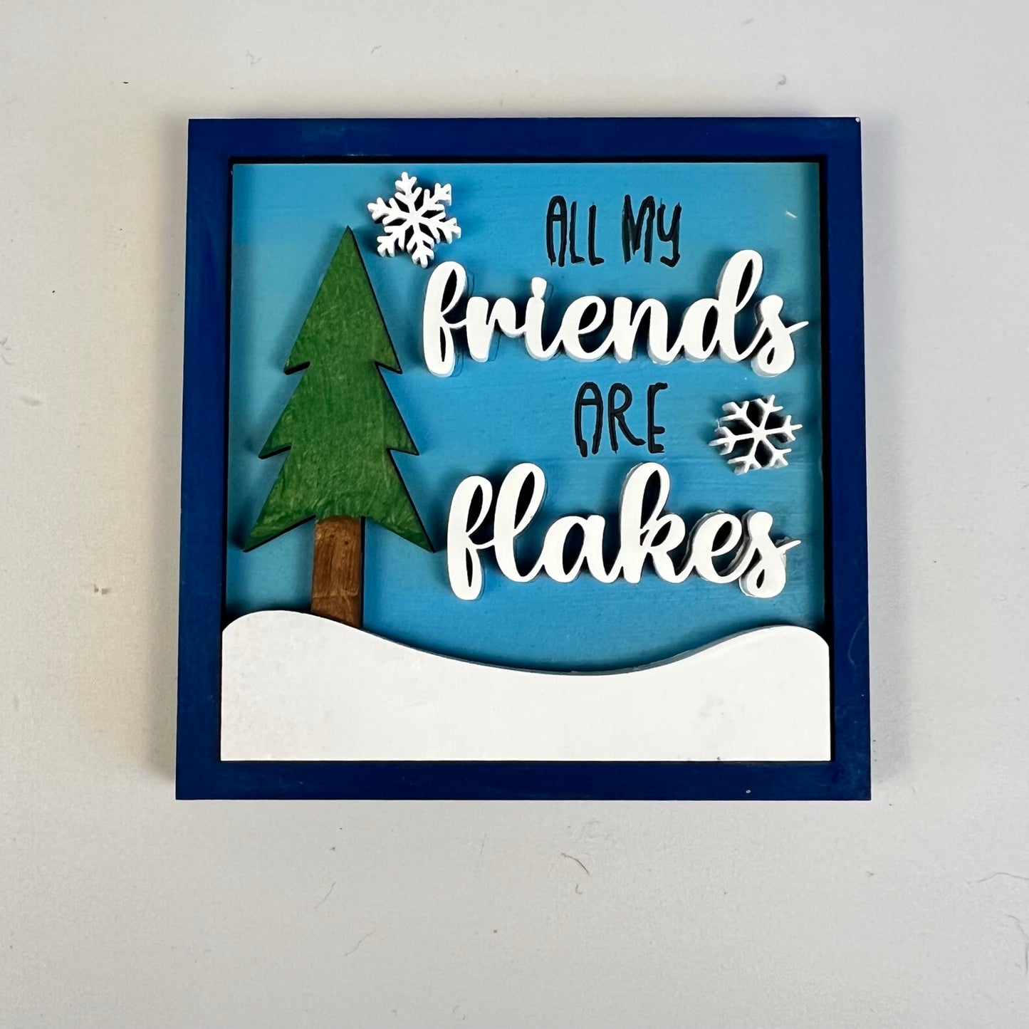 A charming addition to your Winter Interchangeable Squares collection from Janet's Craft Corner, this decorative mini sign features a blue border framing a green tree and snowflakes on a snowy backdrop. Ideal for tiered trays, it includes the playful white text "All my friends are flakes," adding whimsical charm to your decor.