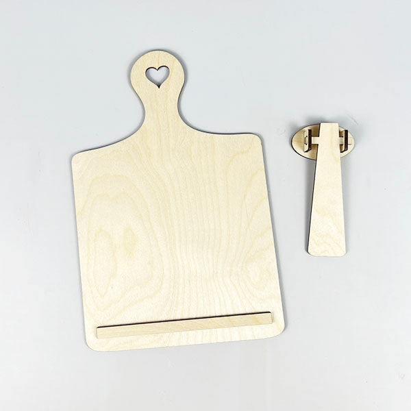 A delightful Breadboard Interchangeable Squares Holder from Janet's Craft Corner, perfect for home décor, lies disassembled on a white surface. With a heart-shaped hole in its handle, the main board features a bottom ledge and sits beside its support piece, reminiscent of an inviting breadboard sign holder.