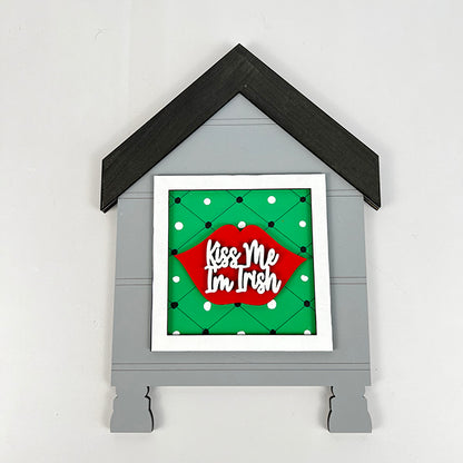 Introducing the House Interchangeable Squares Holder from Janet's Craft Corner, a delightful farmhouse décor piece shaped like a house. It features a charming green polka-dotted square with red lips and the playful message "Kiss Me I'm Irish," adding a whimsical touch to your home. The décor showcases a black roof with a gray house design.
