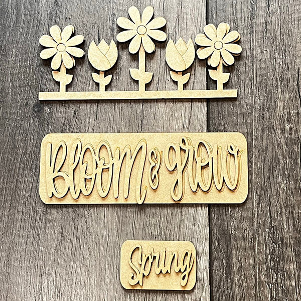 On a gray background, five hand-painted wooden flower cutouts are aligned in a row, captioned with the phrase "bloom & grow" in script. Below, a smaller cursive piece displays "Spring." This delightful DIY home décor kit, known as the Spring Blooms Interchangeable Insert by Janet's Craft Corner, allows for seasonal customization.